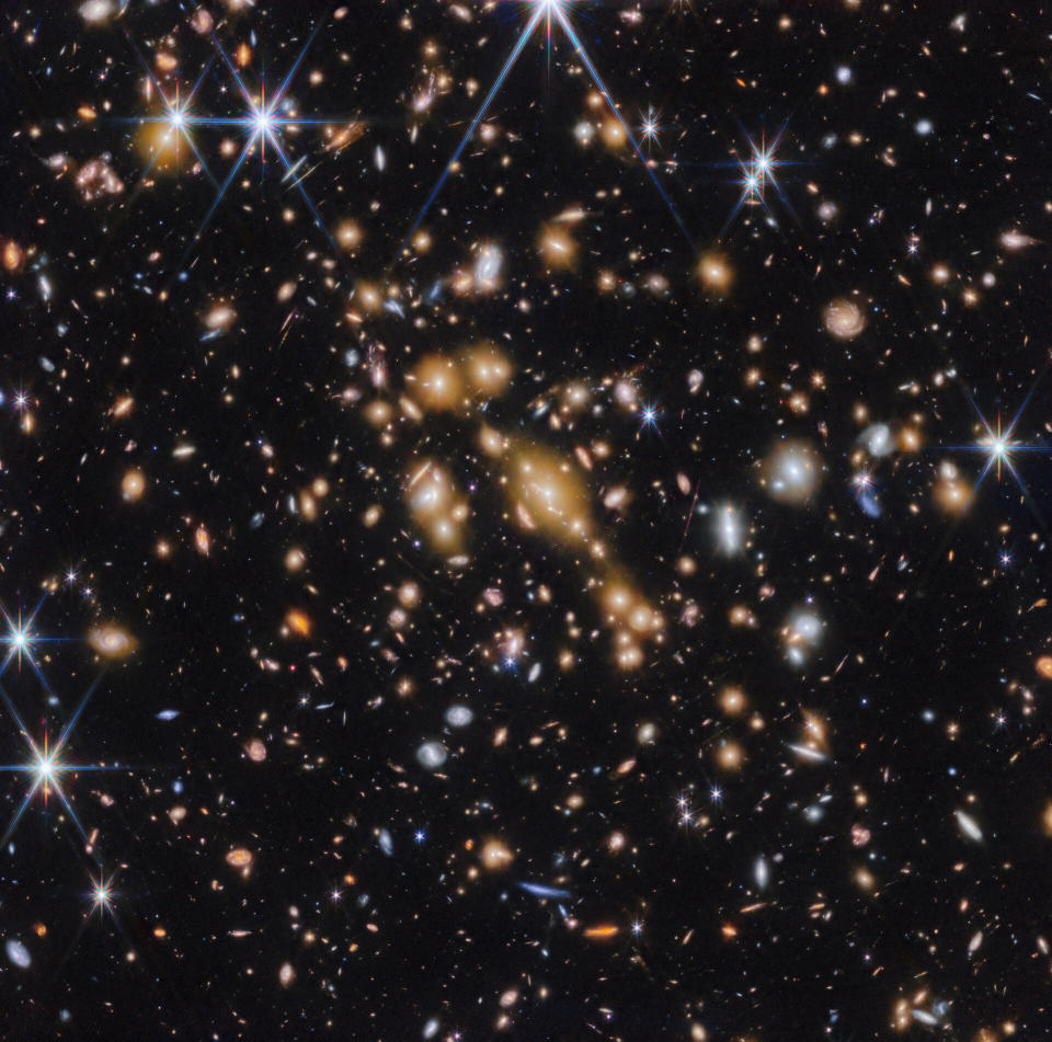 A field of galaxies on the black background of space. In the middle is a collection of dozens of yellowish galaxies that form a foreground galaxy cluster. Among them are distorted linear features, which mostly appear to follow invisible concentric circles curving around the centre of the image. The linear features are created when the light of a background galaxy is bent and magnified through gravitational lensing. A variety of brightly coloured, red and blue galaxies of various shapes are scattered across the image, making it feel densely populated