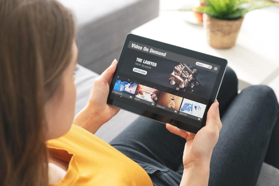 A person seated on their couch while using video-on-demand streaming services on their tablet.