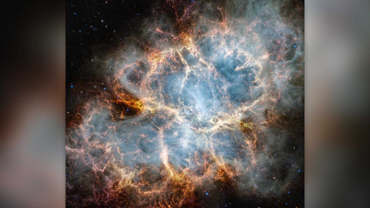  The Crab Nebula. An oval nebula with complex structure against a black background. On the nebula’s exterior, particularly at the top left and bottom left, lie curtains of glowing red and orange fluffy material. Its interior shell shows large-scale loops of mottled filaments of yellow-white and green, studded with clumps and knots. 