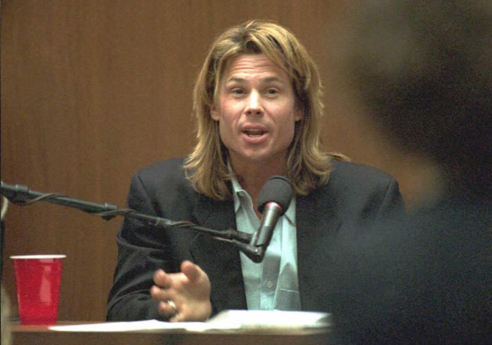 kato kaelin speaks while sitting behind a microphone, he wears a suit jacket and green collared shirt