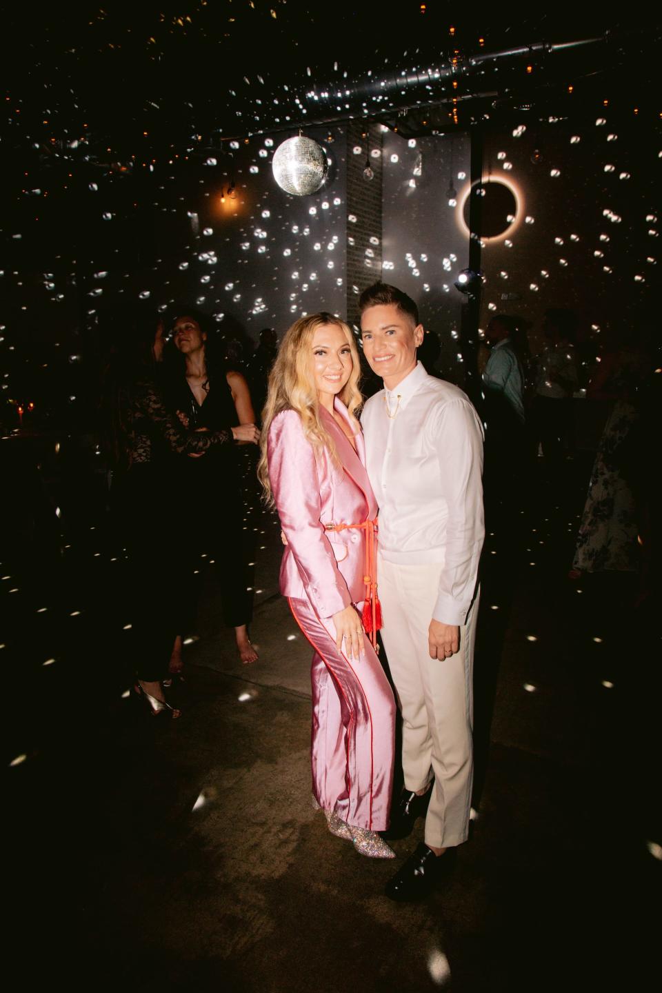 A bride in a pink jumpsuit and a bride in a white suit pose for a photo under a disco ball.