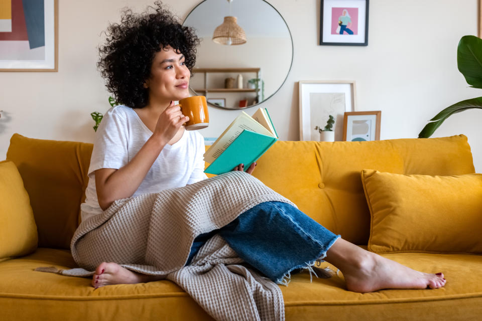Pensive relaxed African american woman reading a book at home, drinking coffee sitting on the couch. Copy space. Lifestyle concept.