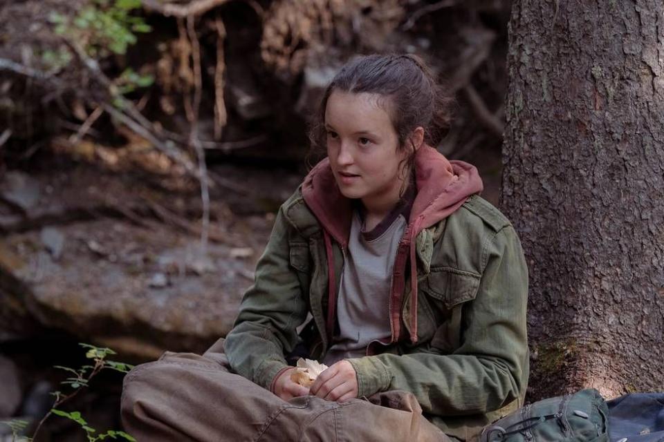 a publicity photo from the last of us featuring bella ramsey sitting down, leaning against a tree, her hands folded, wearing a green jacket