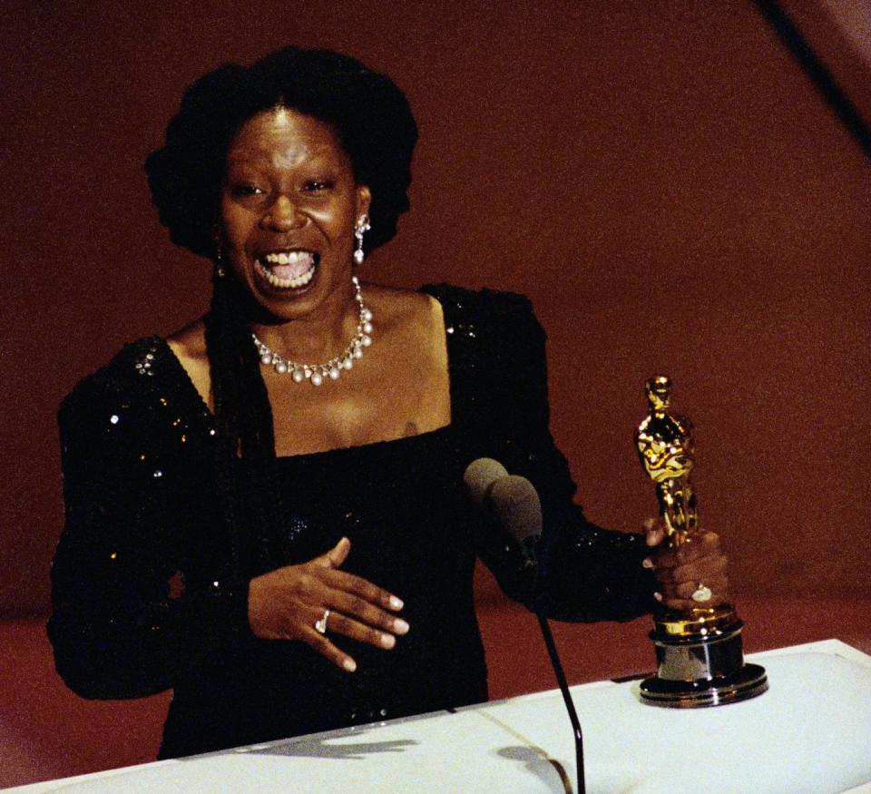 whoopi goldberg wears a black sparkly dress and holds an oscar as she accepts an award for best supporting actress, she is speaking into a microphone at a white podium with the statue in her left hand