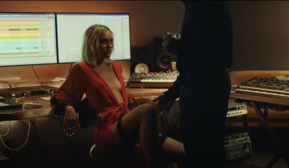 Lily-Rose's character sits in a chair in a music studio and looks up while a man standing touches her knee