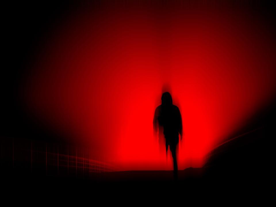 A dark, horror concept. Of a blurred hooded figure silhouetted against lights at night. With a black, red moody edit. - stock photo