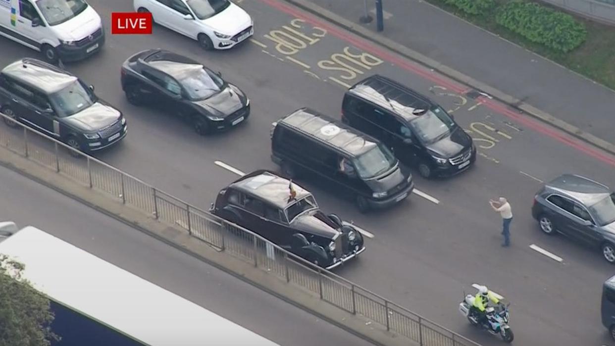 Screengrab from BBC News of a member of the public stepping out into the road towards a car carrying King Charles III and the Queen Consort to take a photo with close protection officers leaving a car which is providing security and gesturing to the member of public