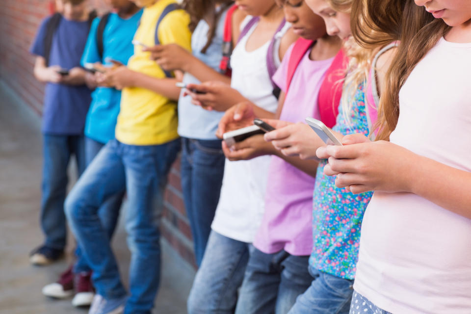 Group of children, standing against a wall, looking down at their smartphones. They are wearing casual clothing