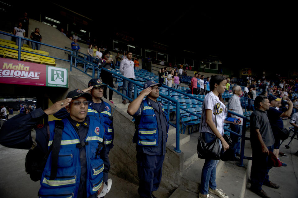 In this Oct. 12, 2018 photo, fire fighters salute during the national anthem at the start of the baseball season's opening game between Leones de Caracas and Tiburones de la Guaira in Caracas, Venezuela. Baseball stadiums have served as a sporting sanctuary where fans of all classes and political backgrounds can set aside their differences and mounting hardships, said Ramon Guillermo Aveledo, a former Venezuela baseball league president and prominent opposition leader. (AP Photo/Fernando Llano)