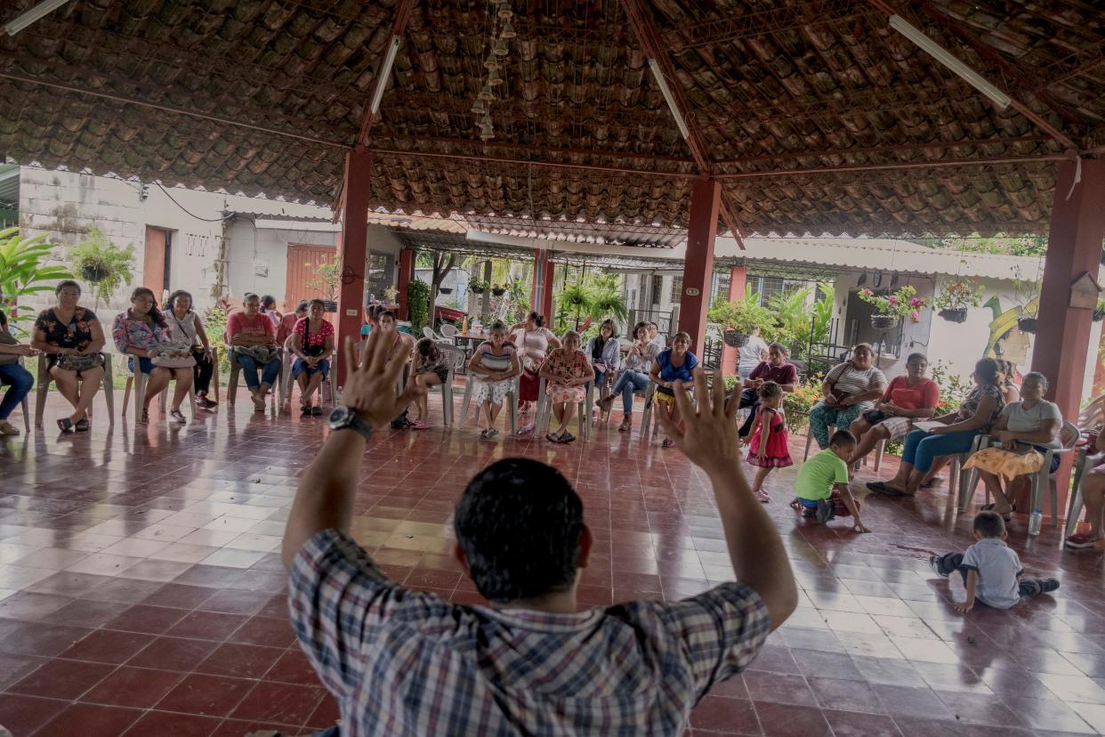 A group of adults sit in a circle, as a man stands at the front with his hands raised.