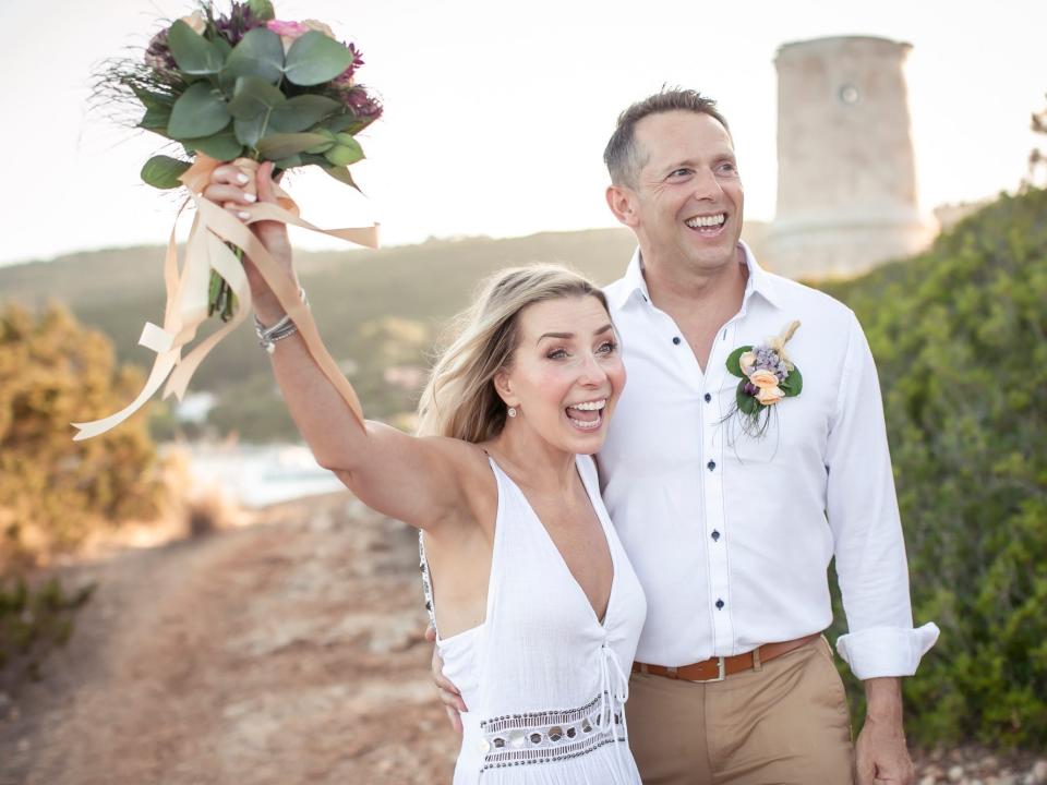 A bride and groom stand together on their wedding day. The bride raises her bouquet in triumph while the groom smiles.