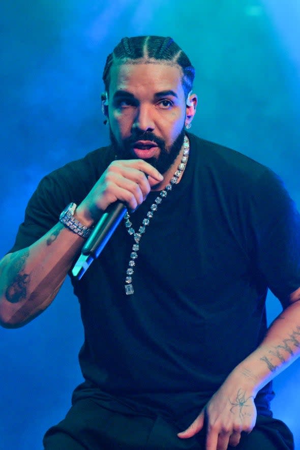 Drake performs on stage wearing a black t-shirt, black pants, and a chain necklace, holding a microphone