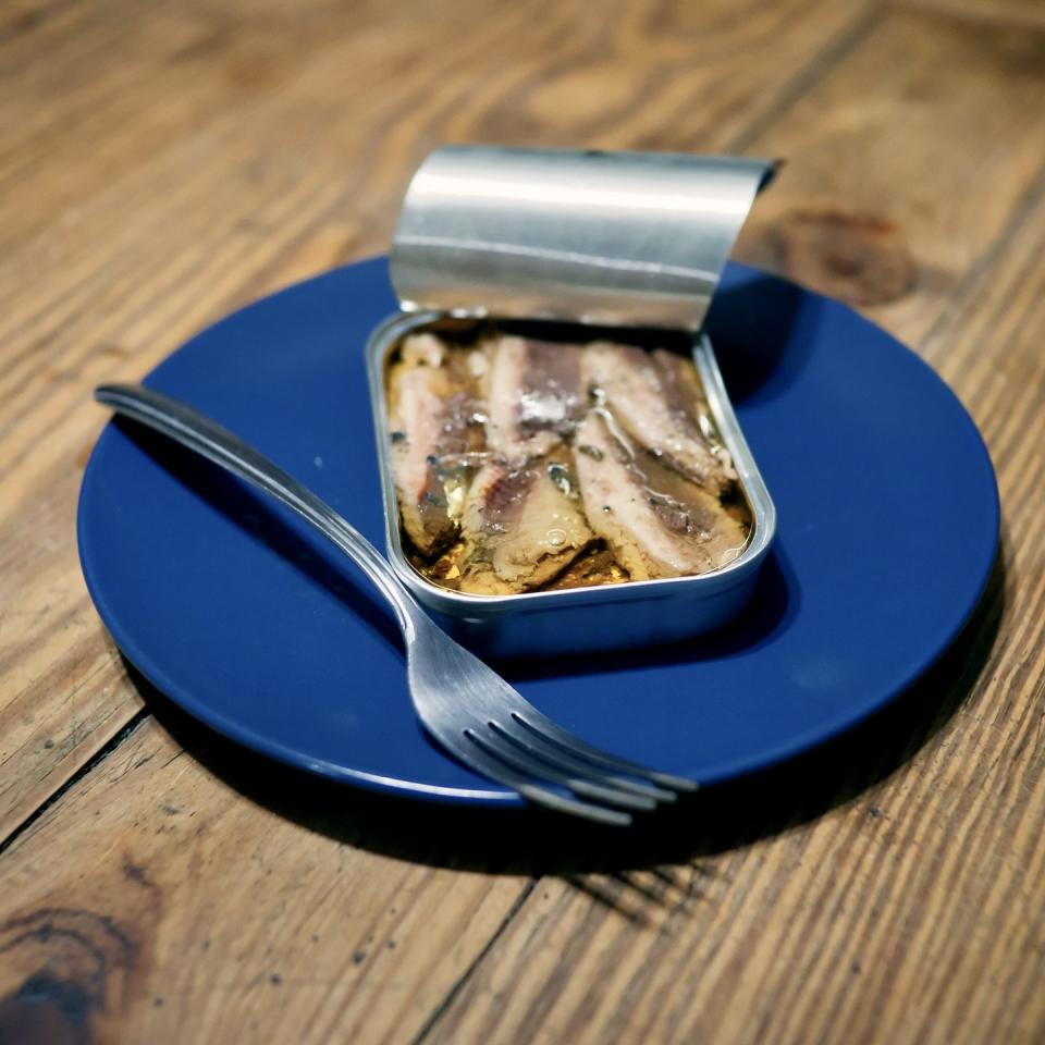 a metal can of sardines provides a satisfactory lunch while plated on a blue saucer with a silver fork on a natural wooden background