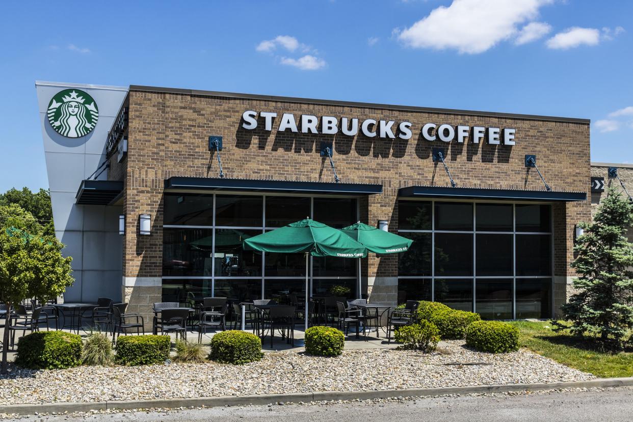 Front exterior of a Starbucks Retail Coffee Store in Indianapolis, Indiana, showing outdoor seating area and surrounding garden, against a blue sky on a sunny day during summer