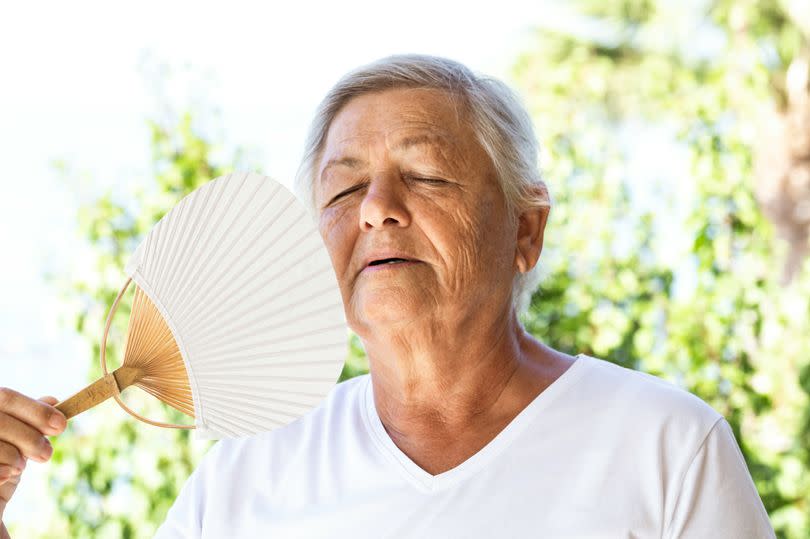 Senior woman is trying to cool herself using hand fan in tropical environment. Representing heat wave solutions.