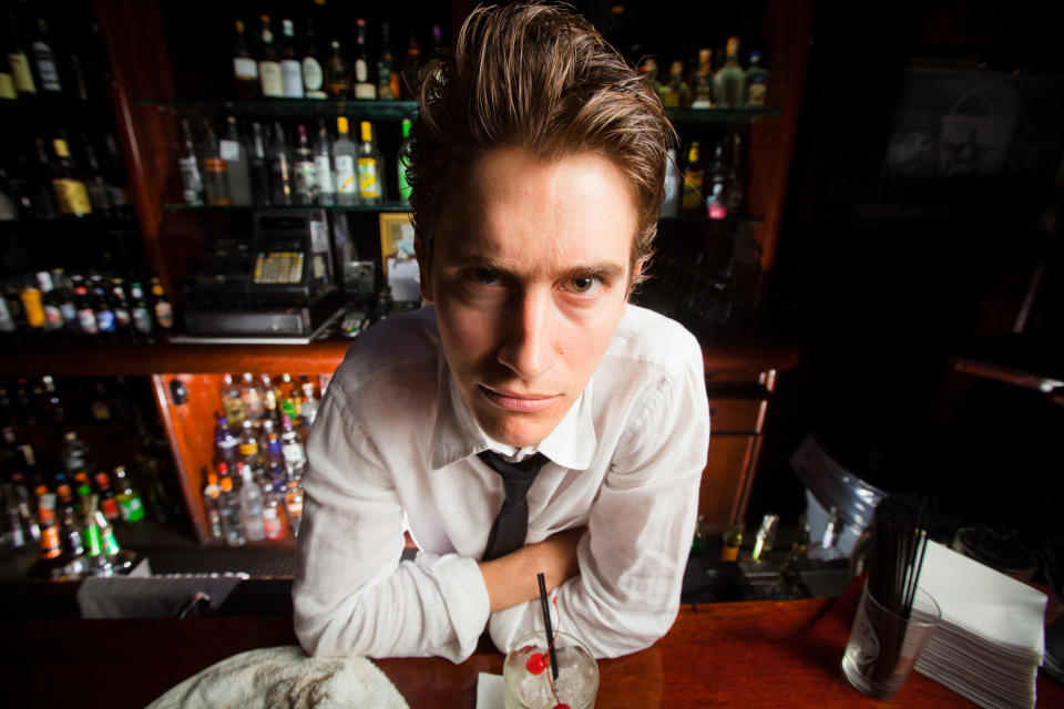 Man in white shirt behind bar with cocktail, looking at camera, exemplifying service industry work