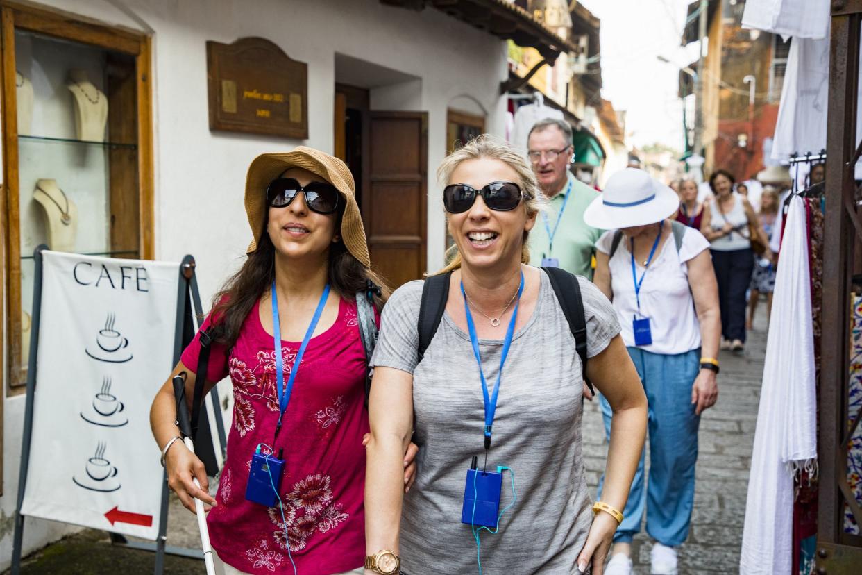 A front-view shot of a multi-ethnic group of tourists walking down a narrow street in Kerala, India. Some of the tourists are visually impaired and can be seen using a blind person's cane and holding onto a friends arm for guidance.