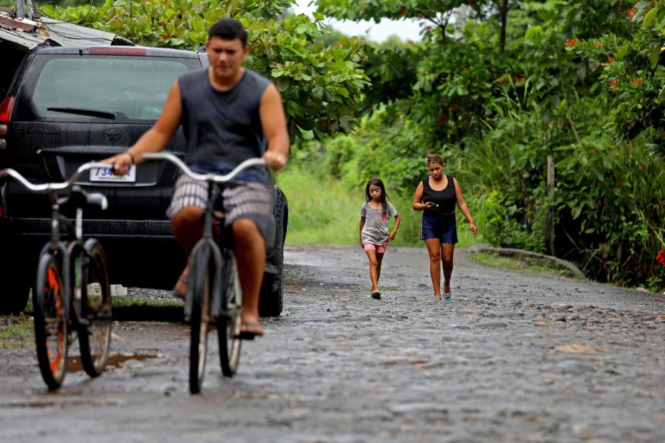 A man riding a bicycle holds onto another bike as a girl and a woman walk behind him