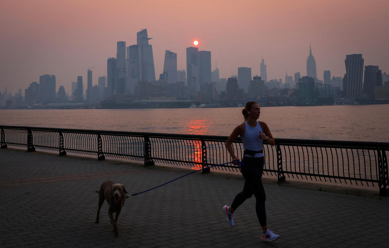 A woman jogs with a dog along the Hudson River, with the air murky and the sun seen dark orange against the high-rises in the distance.