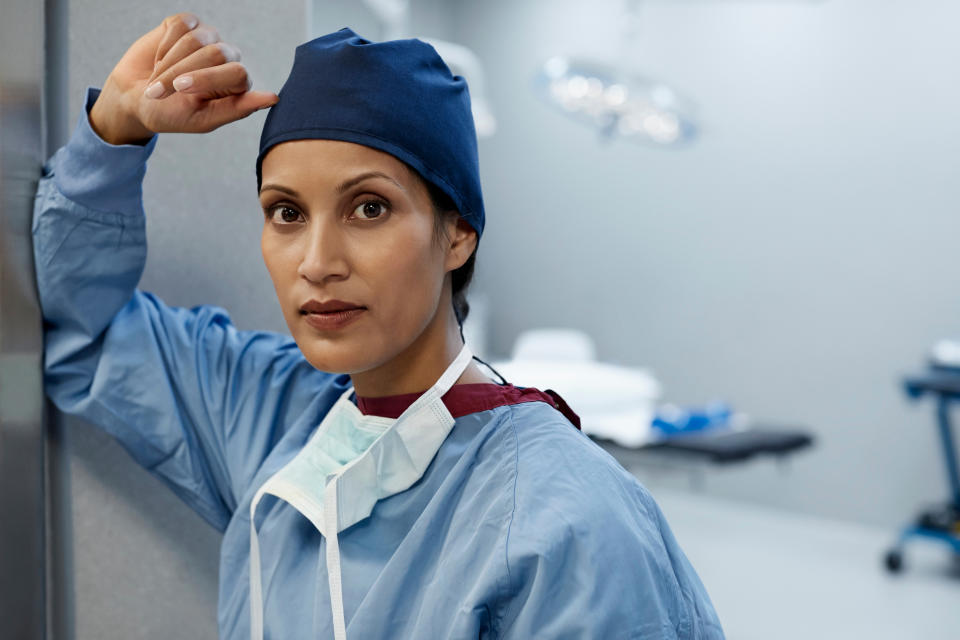 female nurse wearing scrubs leaning against a door frame, looking in to the camera