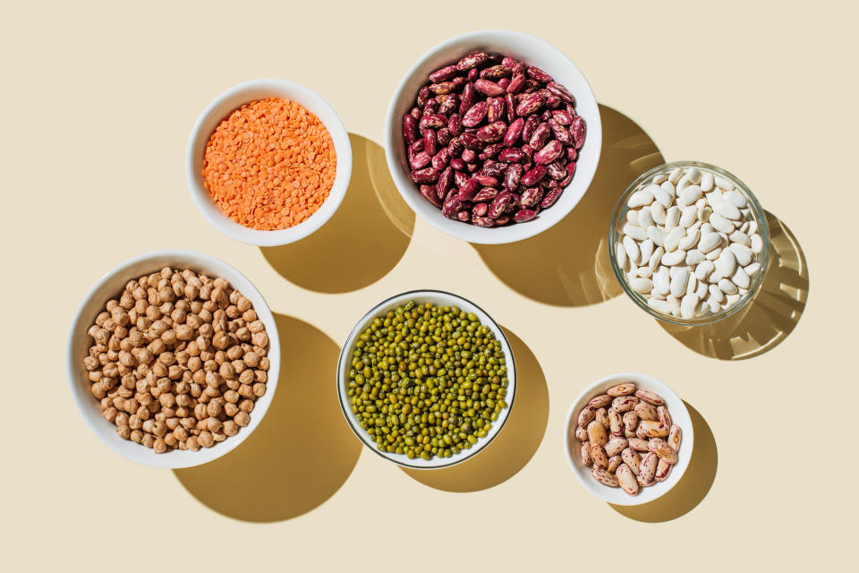 Aerial view of seven white bowls containing various dry beans and lentils, including red lentils, red beans, white beans, chickpeas, mung beans, and pinto beans