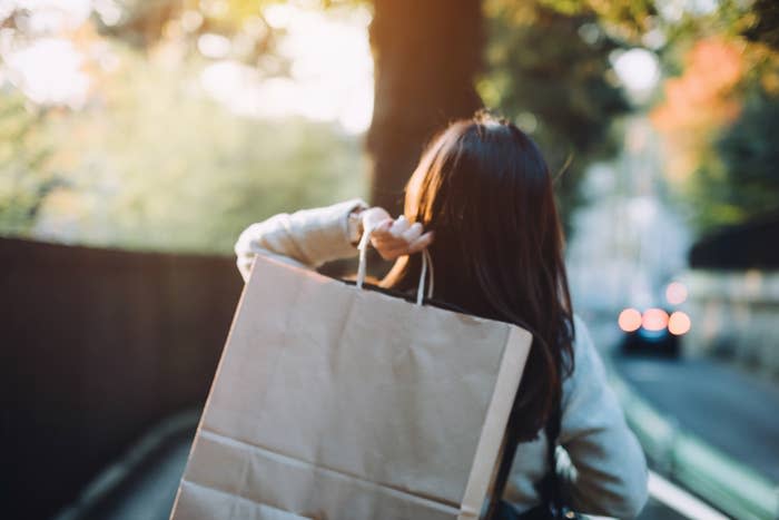 A person with long hair walks along a street carrying a large paper shopping bag on their shoulder. The background appears slightly blurred with evening lights