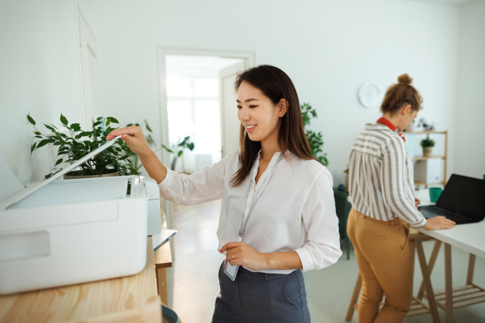 Two women working in an office; one is using a printer while the other works on a laptop