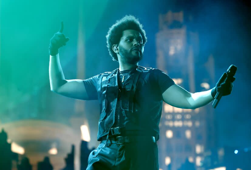 man, the weeknd, with frizzed afro, performing on sofi stadium stage, wearing tactical vest and black shirt, pants and gloves