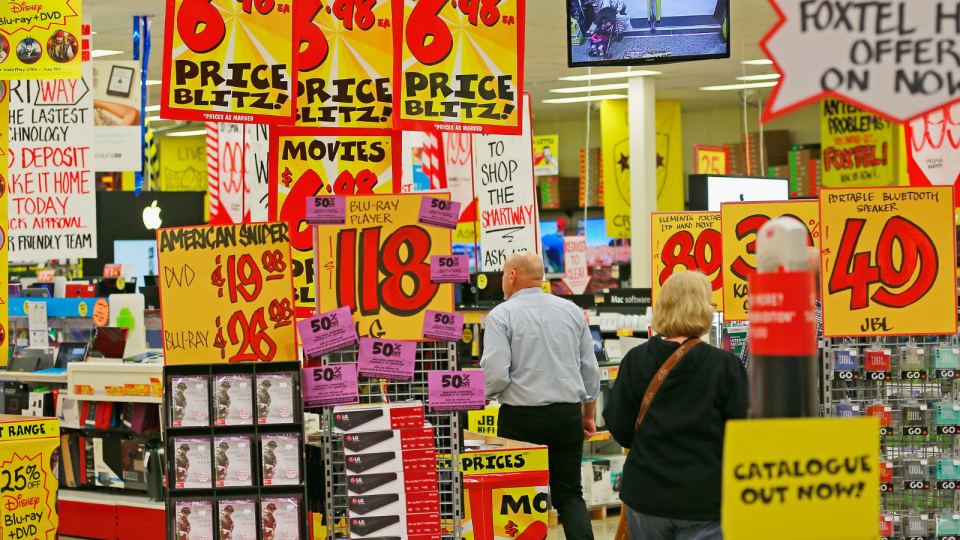 People shopping in JB HIFI amongst bright yellow signs advertising the prices of products