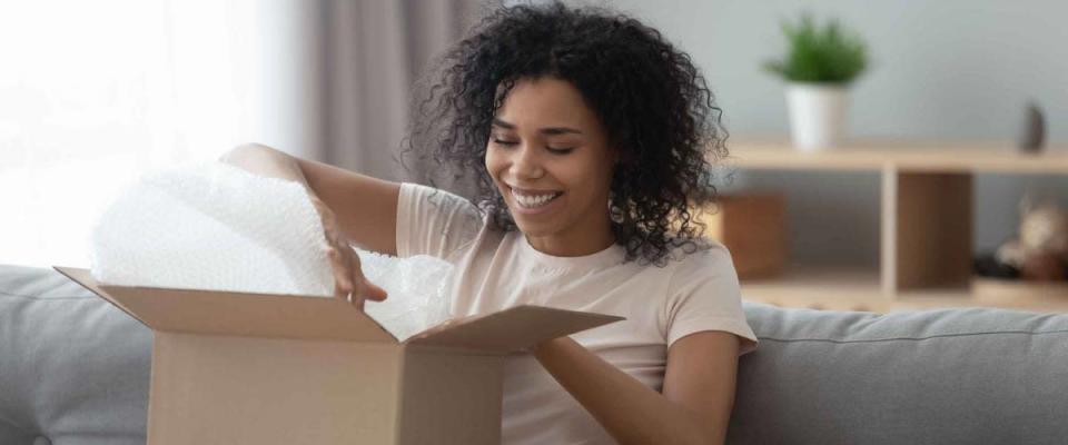 Happy young african woman satisfied customer open parcel cardboard box sit on sofa at home