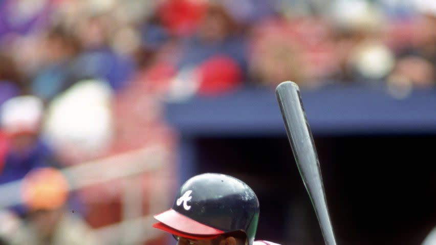 deion sanders holds a bat as he looks forward preparing to hit, he wears a light gray atlanta braves uniform with a blue hitting cap and red and black gloves