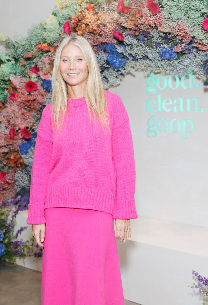 Gwyneth smiling in a pink sweater and skirt, standing in front of a floral backdrop with the words 'Good Clean Goop'