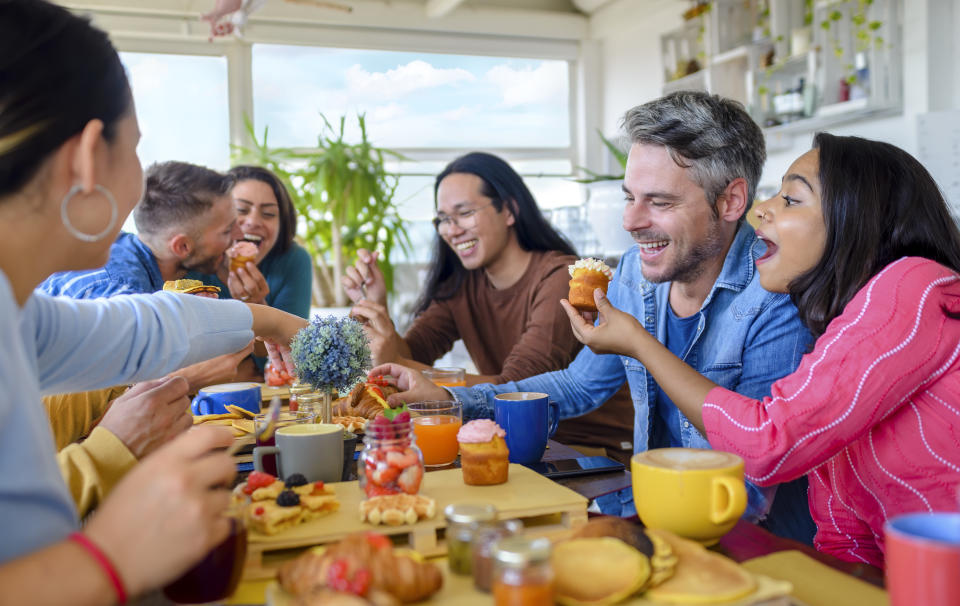 multiethnic group of friends sitting on a table in a bar restaurant eating a muffin making faces. diverse people celerating sweet breakfast together enjoying happy holidays. lifestyle and joy concept