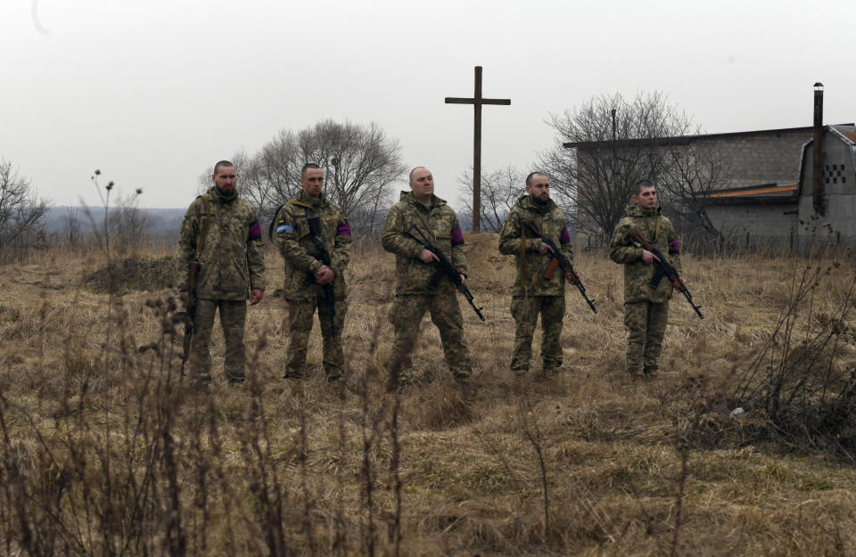 Ukrainian military personnel stand in a field with a cross in the background.