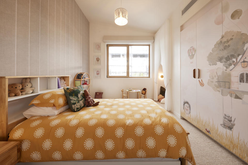 A shot of the bed with a yellow and white doona cover, a window on the far wall and a treehouse mural on the wallpaper. 