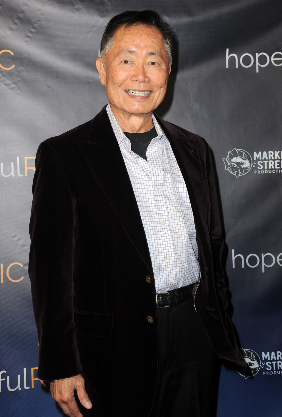 George Takei on the red carpet, smiling and wearing a checkered shirt with a dark blazer at a film event