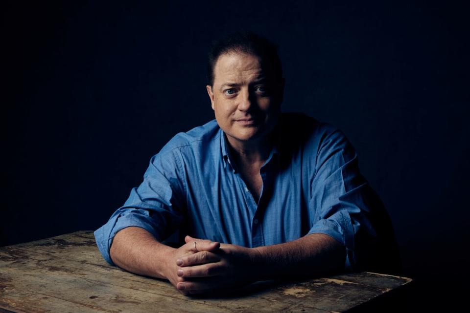 A man wears a dress shirt with the sleeves rolled up as he rests his arms on a table for a portrait.