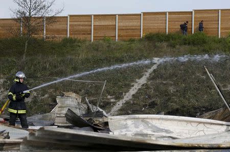 A French fireman extinguish shelters as migrants walk past the camp the day after a fire destroyed large swathes of the Grande-Synthe migrant camp near Dunkirk in northern France April 11, 2017 following skirmishes on Monday that injured several people. REUTERS/Pascal Rossignol