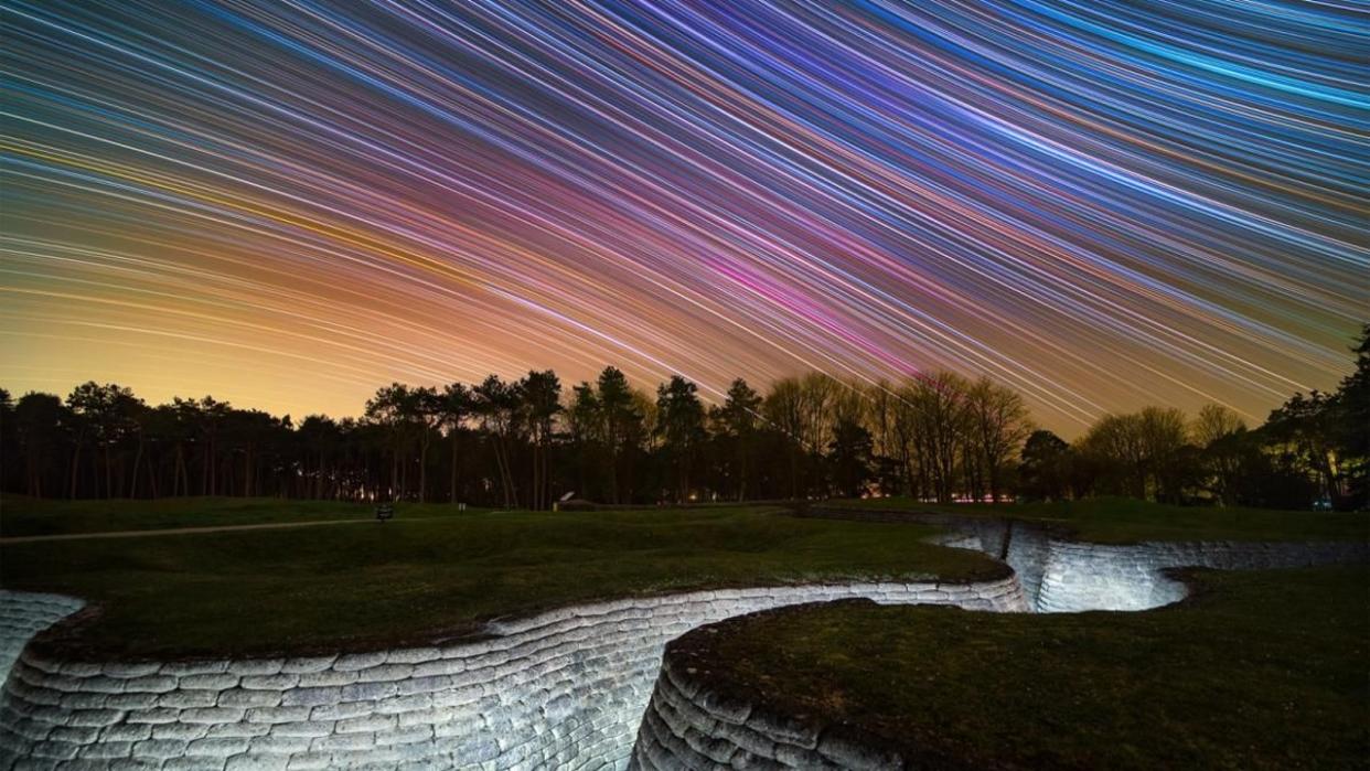  A white-bricked trench lays recessed in the pale green grass of a World War I memorial in France. Above, the night sky is diagonally streaked with a rainbow of stars. 