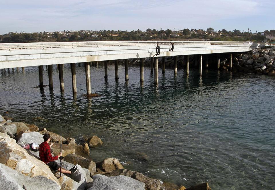 FILE - This Jan. 7, 2014 file photo shows a fisherman, left, enjoys the unseasonably warm Southern California weather as two teenage boys leap from a Highway 101 overpass into the San Batiquitos Lagoon inlet in Carlsbad, Calif. For a chilly and snowbound East, here’s a dose of cold reality: The nation’s temperature in January was pretty normal because a warm West offset a cool East. The National Oceanic Atmospheric Administration said January in the Lower 48 states was the 53rd coldest of 120 years. The nation averaged only one tenth of a degree below normal for January. (AP Photo/Lenny Ignelzi, File)