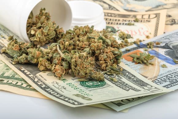 A tipped-over bottle of dried cannabis flower lying atop a messy pile of cash bills.