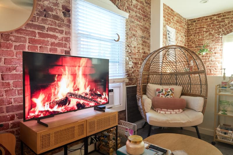 Brick-walled living room with woven chair and TV on media stand.
