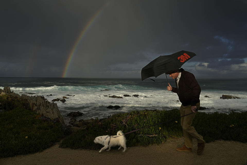 FILE - A man huddles under an umbrella as he walks a dog along the shoreline at Pacific Grove, Calif., on Feb. 2, 2024. California's current rainy season got off to a slow start but has rebounded with recent storms that have covered mountains in snow and unleashed downpours, flooding and mudslides. The water content of the vital Sierra Nevada snowpack has topped 80% of normal to date while downtown Los Angeles has already received more than an entire year's average annual rainfall. (AP Photo/Rich Pedroncelli, File)