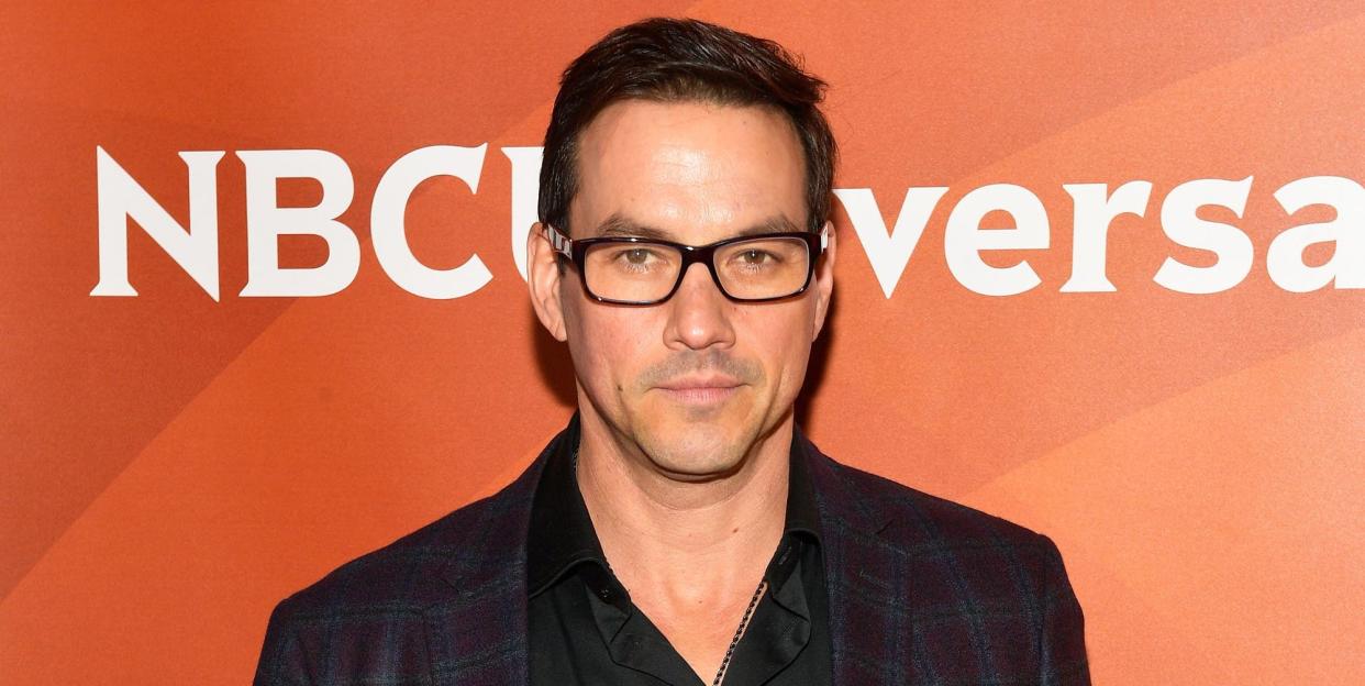 general hospital actor tyler christopher with short black hair and glasses, in a checked jacket, black shirt and jeans against an orrange nbc universal background