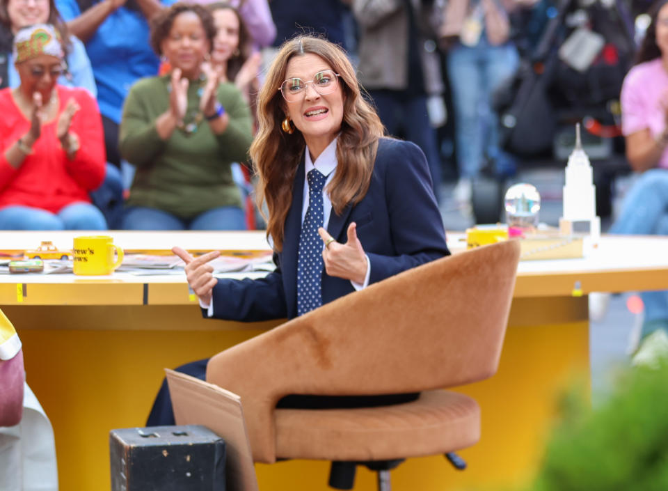 Woman in a suit and polka dot tie sits at a desk outdoors, looking off-camera, pointing with both hands, and smiling. Audience clapping in the background