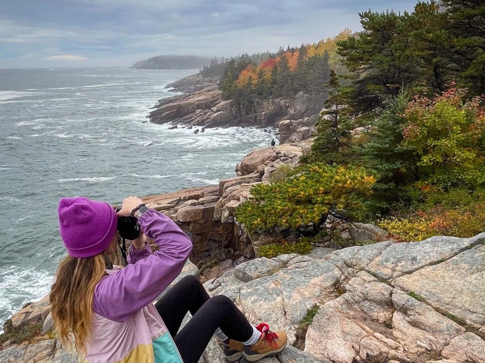 Emily sits on a rock by the water and takes photos of the fall foliage. Her backpack, covered in patches, sits next to her on the rock. 