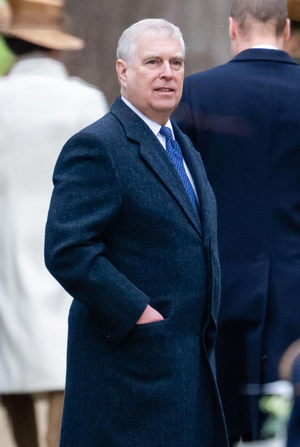 prince andrew stands with his hand in his coat pocket, he wears a white collared shirt and blue patterned tie and looks right