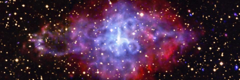 swirling diamond shaped cloud of blue dust with purple and much red along the edges in space with a bright spot in the center and numerous distant galaxies in the background