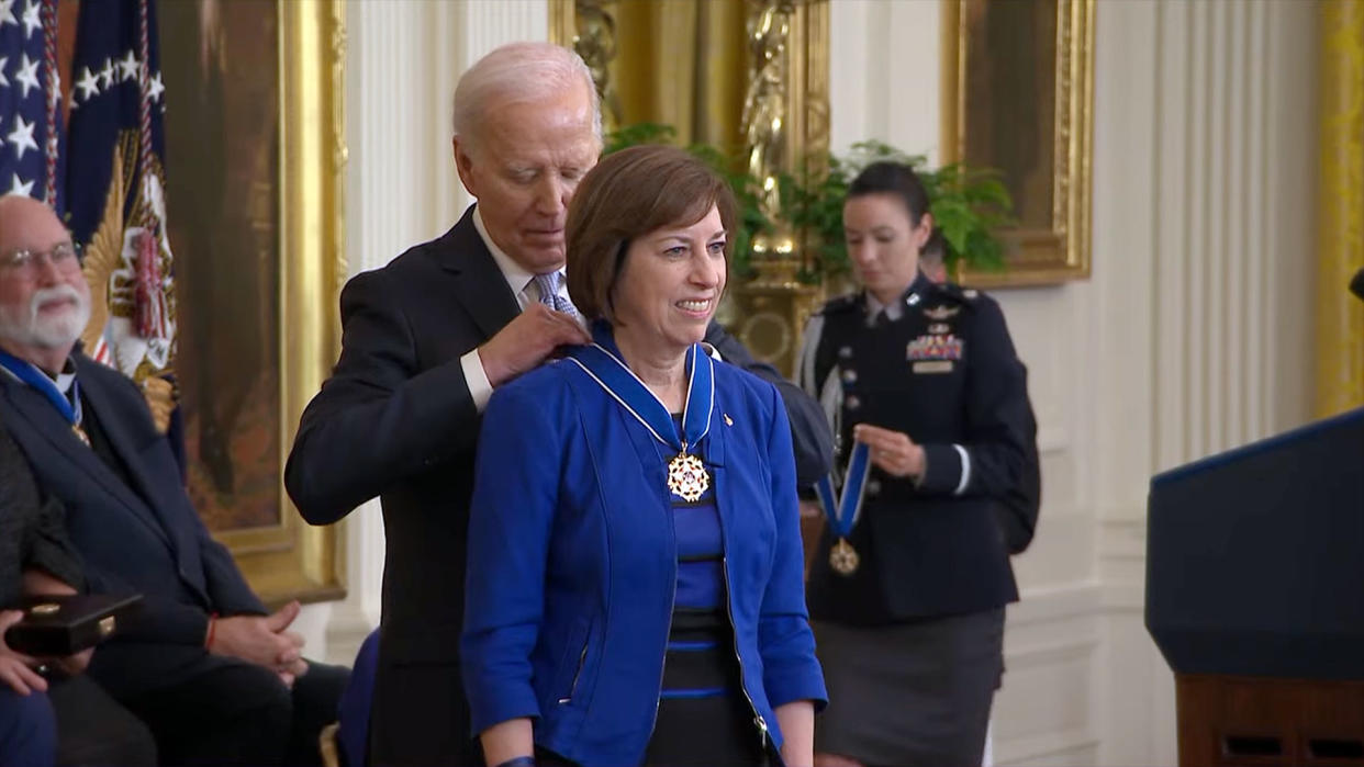  An old man wearing a suit drapes a medal around the neck of a smiling woman in a blue dress. 