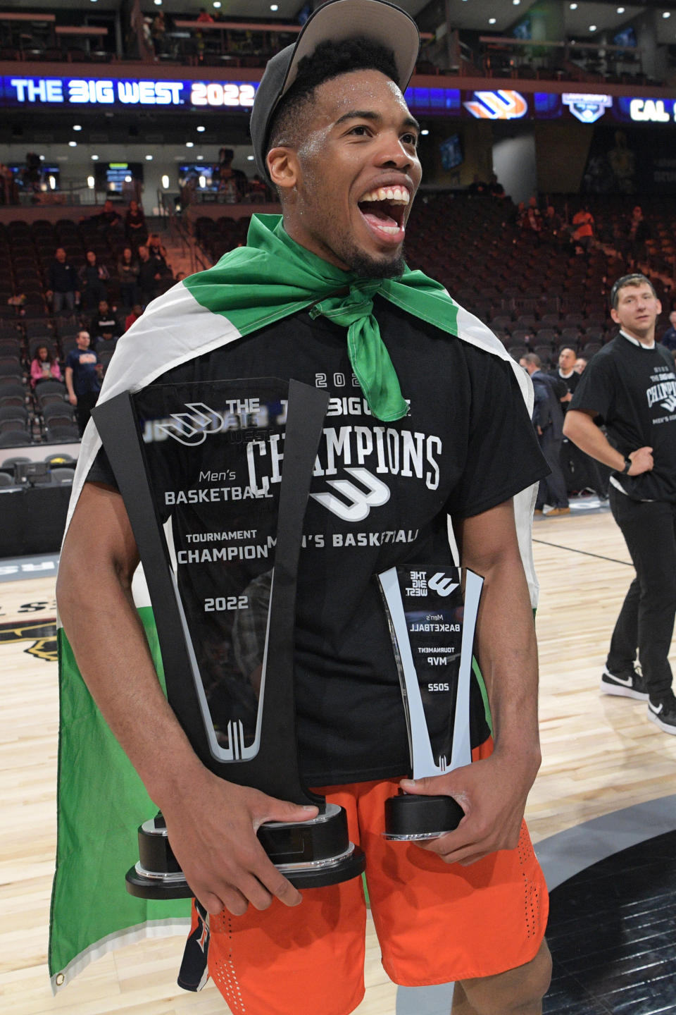 E.J. Anosike poses while smiling, holding two trophies, and wearing a Nigerian flag draped around him as a cape.